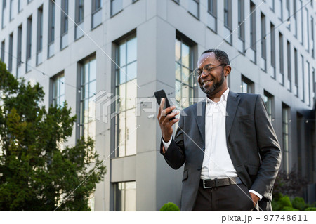 Successful and satisfied african american businessman using phone outside modern office building, mature man in business suit smiling and looking at smartphone screen texting. Successful and satisfied african american businessman using phone outside modern office building, mature man in business suit smiling and looking at smartphone screen texting. 97748611