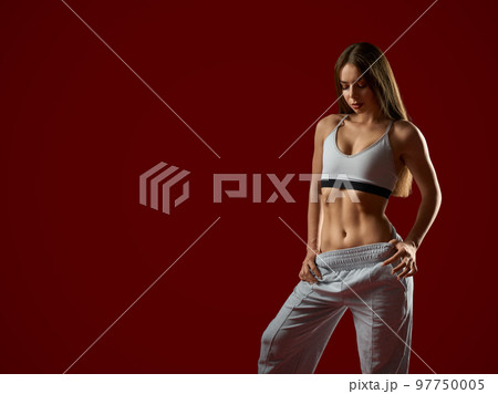 Front view of pretty brunette female model with press cubes in white sport wear posing on red studio background, looking down. Sexy athletic fit woman trainer standing posing, showing stomach. 97750005