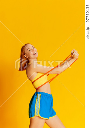 Portrait of young redhead girl in sportswear training with sports equipment over yellow background. Hands exercises with expander 97750133