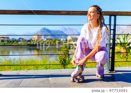 Photo of cheerful young happy woman wear white t-shirt and bell-bottomed trousers with pink pastel color roller skates outdoors in sunny day 97753091