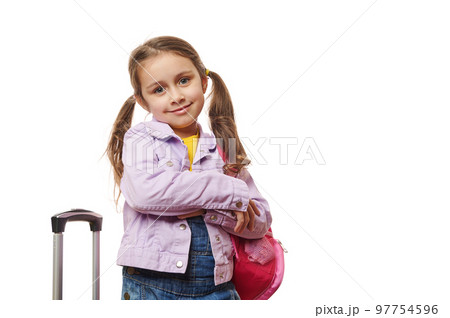 Caucasian preschooler lovely little traveler girl with two ponytails, dressed in purple jacket and blue denim overalls, smiling at camera, posing with pink backpack and suitcase on white background. 97754596