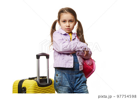 Isolated portrait on white background of a little child girl with two ponytails, dressed in purple jacket, posing with her arms crossed, standing near yellow suitcase over white background. Copy Space Isolated portrait on white background of a little child girl with two ponytails, dressed in purple jacket, posing with her arms crossed, standing near yellow suitcase over white background. Copy Space 97754598