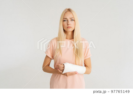Studio portrait of unhappy injured blonde young woman with broken arm wrapped in plaster bandage looking at camera, standing on white isolated background. Concept of insurance and healthcare. 97765149