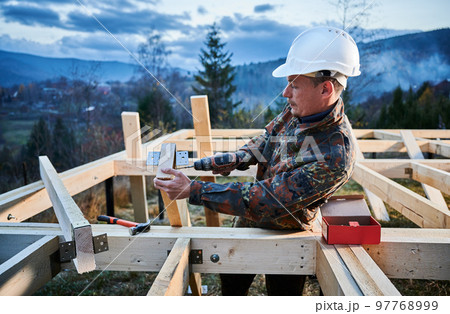 Man worker building wooden frame house on pile foundation. Carpenter installing furniture for wooden truss, using hammer and screwdriver. Carpentry concept. 97768999