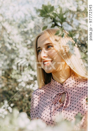 Blonde girl on a spring walk in the garden with cherry blossoms. Female portrait, close-up. A girl in a pink polka dot dress. 97772545
