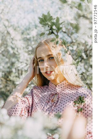 Blonde girl on a spring walk in the garden with cherry blossoms. Female portrait, close-up. A girl in a pink polka dot dress. 97772546