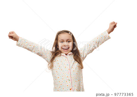 Cheerful baby girl in white pyjama with colorful dots, raising arms up, rejoicing, expressing amazement and happiness, smiling looking at camera, on white background. Space for your advertising text Cheerful baby girl in white pyjama with colorful dots, raising arms up, rejoicing, expressing amazement and happiness, smiling looking at camera, on white background. Space for your advertising text 97773365
