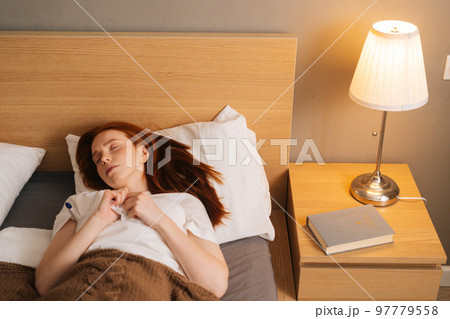 Portrait of sick tired young woman with flu lying under blanket with closed eyes on bed holding thermometer in armpit. Ill female with cold lying in bed with high fever in bedroom at home. 97779558