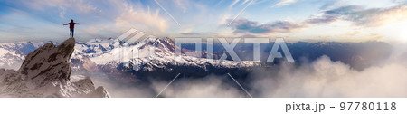 Adventurous Woman Hiker standing on top of icy peak with rocky mountains in background. 97780118