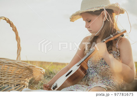 Portrait of little girl in hat sitting on blanket in dry hay field, having picnic, learning playing ukulele. Side view 97781675