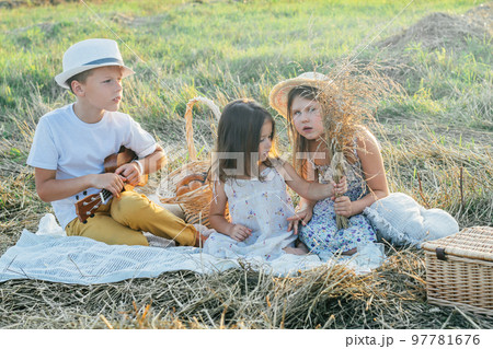 Portrait of glad three kids sitting on blanket in field, playing ukulele, having picnic with drinks and tasty food. 97781676