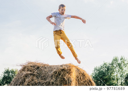 Portrait of barefoot boy in white short and yellow trousers jumping on haystack in field. Outdoor activity. Side view Portrait of barefoot boy in white short and yellow trousers jumping on haystack in field. Outdoor activity. Side view 97781689