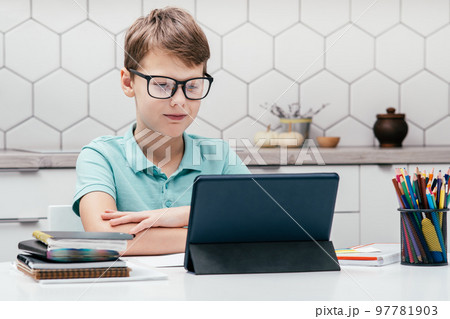 Portrait of young preteen calm boy wearing glasses, sitting at desk, looking at screen of tablet, having online lesson. 97781903