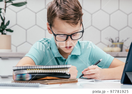 Portrait of preteen focused boy sitting at desk near notebooks, tablet, writing on notebook, holding pen with left hand. Portrait of preteen focused boy sitting at desk near notebooks, tablet, writing on notebook, holding pen with left hand. 97781925