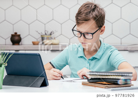 Portrait of preteen serious boy sitting at desk, looking at screen of tablet, doing homework, writing with left hand. 97781926