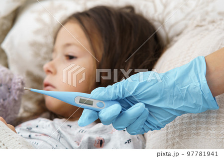 Cropped doctor hand in glove hold electronic thermometer against calm, sad, silent little girl in bed. Quarantine time Cropped doctor hand in glove hold electronic thermometer against calm, sad, silent little girl in bed. Quarantine time 97781941