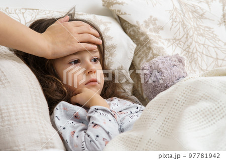 Unrecognizable woman hand touching little girl forehead in bedroom. Bored girl lying with bear toy in white soft bed Unrecognizable woman hand touching little girl forehead in bedroom. Bored girl lying with bear toy in white soft bed 97781942