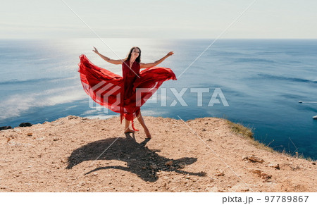 Woman in red dress on sea. Side view a Young beautiful sensual woman in a red long dress posing on a rock high above the sea on sunset. Girl on the nature on blue sky background. Fashion photo. 97789867