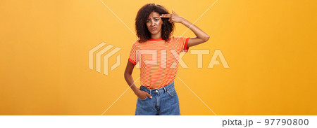 Portrait of displeased gloomy and sad annoyed dark-skinned female with curly hairstyle showing gun with hand near temple frowning wanting to kill herself from exhaust and boredom over orange wall Portrait of displeased gloomy and sad annoyed dark-skinned female with curly hairstyle showing gun with hand near temple frowning wanting to kill herself from exhaust and boredom over orange wall 97790800