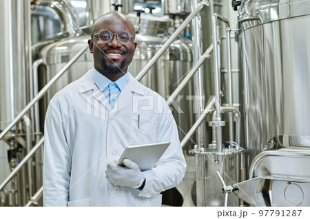 Portrait of successful engineer with digital tablet smiling at camera standing in workshop with big barrels in background Portrait of successful engineer with digital tablet smiling at camera standing in workshop with big barrels in background 97791287