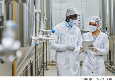 Two professional engineers in white coats and masks discussing something on digital tablet in workshop of brewery Two professional engineers in white coats and masks discussing something on digital tablet in workshop of brewery 97791336