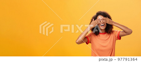 Closing my eyes and counting ten. Portrait of charming dreamy and happy funny african american curly-haired female in striped trendy t-shirt covering sight with palm and smiling waiting surprise 97791364