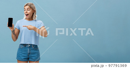 Look at this awesome smartphone. Impressed enthusiastic good-looking female blonde with cool tattoo on arm dropping jaw from thrill and excitement pointing at device screen over blue background Look at this awesome smartphone. Impressed enthusiastic good-looking female blonde with cool tattoo on arm dropping jaw from thrill and excitement pointing at device screen over blue background 97791369