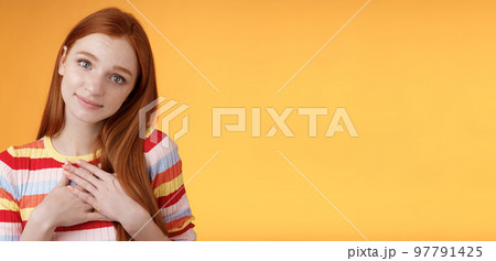 Lovely silly cute redhead young european girl tilting head look gently smiling tender camera press palms heart thankful receive nice gift gazing gratitude happiness, standing orange background 97791425