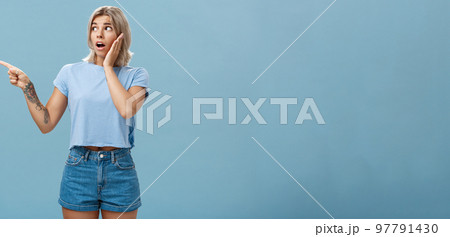 Studio shot of nervous shocked good-looking young woman with fair hair and tatooed arm gasping pointing and looking left with worry holding hand on cheek posing over blue background 97791430