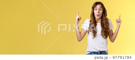Impressed enthusiastic surprised ambushed cute female model fold lips wow admiration joy stare camera fascinated pointing up index fingers top copy space show you stunning offer yellow background 97791784