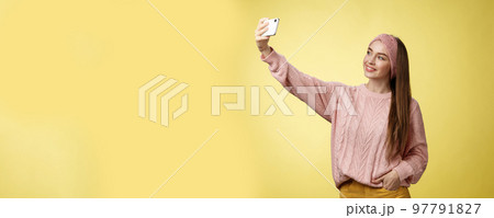 Popular glamour young female internet lifestyle blogger taking selfie on new smartphone extending arm taking picture herself against yellow background smiling at cellphone screen, posing cheeky Popular glamour young female internet lifestyle blogger taking selfie on new smartphone extending arm taking picture herself against yellow background smiling at cellphone screen, posing cheeky 97791827