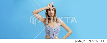 Waist-up shot of stylish self-assured woman with tattoo feeling like winner mocking over losers sticking out tongue and showing letter L over forehead making rebellious expression against blue wall 97792511