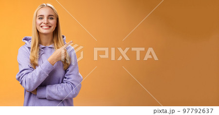 Charismatic modern millennial cute blond woman in hoodie smiling happily discussing cool new promo pointing right recommend check out visit interesting store near corner, orange background 97792637