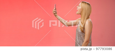 Profile shot funny carefree blond woman hold smartphone raised open mouth wide yelling recording video own shout scream, standing silver glamour dress near red background fool around Profile shot funny carefree blond woman hold smartphone raised open mouth wide yelling recording video own shout scream, standing silver glamour dress near red background fool around 97793086