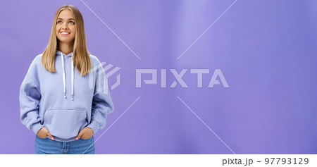 Winter comming. Dreamy kind and cute young girl with straight fair hair and tan wearing hoodie looking at upper left corner with nice white smile and admiring expression holding hand in pockets 97793129