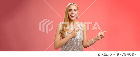 Excited carefree happy young attractive woman joyful attend awesome party in silver beautiful glittering dress pointing left suggesting come together inviting dance smiling cute red background 97794087