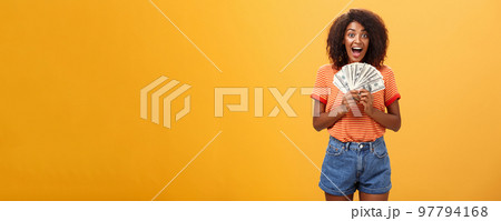 Woman cannot hide happiness winning lottery. Portrait of surprised and happy lucky african american young woman with curly hair holding bunch of money and yelling from delight receiving paycheck Woman cannot hide happiness winning lottery. Portrait of surprised and happy lucky african american young woman with curly hair holding bunch of money and yelling from delight receiving paycheck 97794168