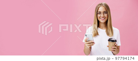 Pleasant friendly-looking girl smiling at camera holding paper cup of coffee and smartphone. Portrait of joyful nice woman drinking morning drink, posing opinion about cafe in internet over pink wall 97794174