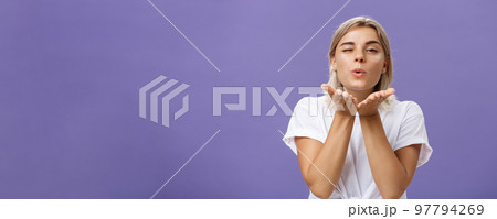 Blowing sweet kiss to all fans. Flirty tender and cute stylish european female with blond hair and tanned skin holding palms near mouth folding lips while sending mwah at camera over purple wall 97794269