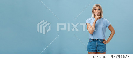 Studio shot of dreamy optimistic good-looking fair-haired female in denim shorts saying shh showing shush gesture with index finger over mouth smiling hiding secret over blue background Studio shot of dreamy optimistic good-looking fair-haired female in denim shorts saying shh showing shush gesture with index finger over mouth smiling hiding secret over blue background 97794623