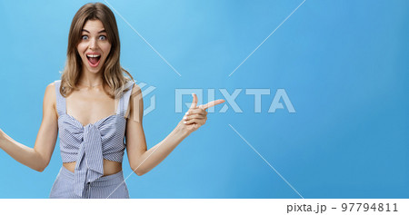 Amazed and surprised charismatic attractive woman raising hands pointing sideways left and right with impressed overwhelmed expression, smiling broadly liking both sides of copy space over blue wall 97794811