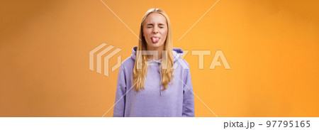 Playful amused charismatic young funny blond girl having fun close eyes showing tongue rebellious fool around unbothered enjoying carefree sunny spring days standing orange background 97795165