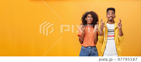 Couple of friends praying and looking up in sky intense crossing fingers for good luck clenching teeth faithfully hoping parents not saw them smoking standing together over orange background 97795587