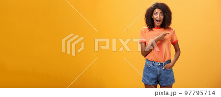 Hey awesme copy space there. Portrait of impressed and surprised enthusiastic young african american female with afro hairstyle pointing right astonished looking energized and curious over orange wall Hey awesme copy space there. Portrait of impressed and surprised enthusiastic young african american female with afro hairstyle pointing right astonished looking energized and curious over orange wall 97795714