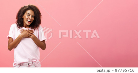 Portrait of charming delighted african american woman with curly haircut holding palms on heart pleased and grateful thanking friend for help smiling happily and thankful at camera over pink wall Portrait of charming delighted african american woman with curly haircut holding palms on heart pleased and grateful thanking friend for help smiling happily and thankful at camera over pink wall 97795716