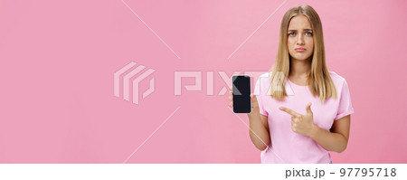 Gloomy and sad cute young female showing friend negative feedback about her project in internet pointing at smartphone screen standing concerned and upset against pink background Gloomy and sad cute young female showing friend negative feedback about her project in internet pointing at smartphone screen standing concerned and upset against pink background 97795718