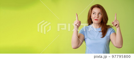 Curious thoughtful cute redhead female middle-aged ginger mother smirking thinking look aside pointing up index fingers deciding choice pondering choosing between variants green background 97795800