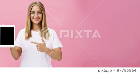 Waist-up shot of optimistic and joyful female showing cool digital tablet pointing at device screen and smiling broadly at camera giving advice what app useful posing in glasses against pink wall 97795964