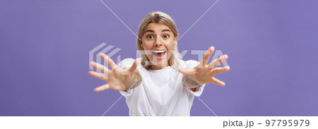Waist-up shot of amused and excited attractive stylish young woman in white t-shirt pulling hands at camera with desire smiling thrilled and happy wanting hug or take something over purple background 97795979