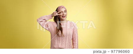 Cheerful happy glamour young european woman in pink knitted sweater, wearing headband, winking flirty and cute, showing victory or pease sign over eye, feeling excited and joyful over yellow wall 97796060